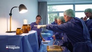 Schülerinnen und Schüler in OP-Kleidung bei einem Workshop in einer Klinik. Eine Person zeigt auf Präparate in braunen Gläsern. © Screenshot 