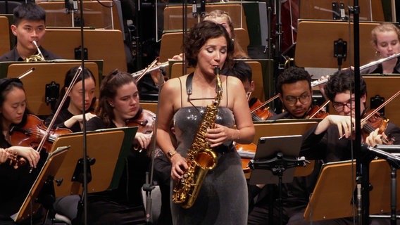 Eine Saxofonistin in einem Konzert. Im Hintergrund das Orchester. © Screenshot 