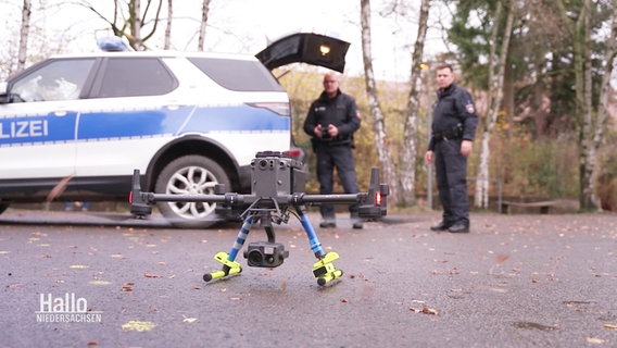 Im Vordergrund steht eine Drohne auf dem Asphalt, im Hintergrund stehen zwei Polizisten neben einem Einsatzwagen. © Screenshot 