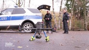 Im Vordergrund steht eine Drohne auf dem Asphalt, im Hintergrund stehen zwei Polizisten neben einem Einsatzwagen. © Screenshot 