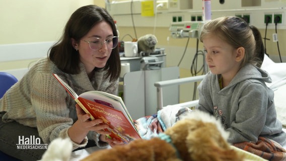 Eine junge Frau zeigt einem kleinen Mädchen etwas in einem Buch. Sie sind in einem Krankenhauszimmer. © Screenshot 