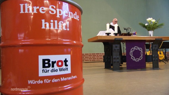 Ein Metallfass in orange. Darauf geschrieben steht "Brot für die Welt". Im Hintergrund ein Pfarrer an einem Altar. © Screenshot 