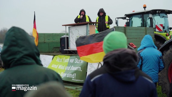 Bei einer Demonstration auf einem Acker hören viele Teilnehmende einer Rednerin auf einem Trecker-Anhänger zu. © Screenshot 