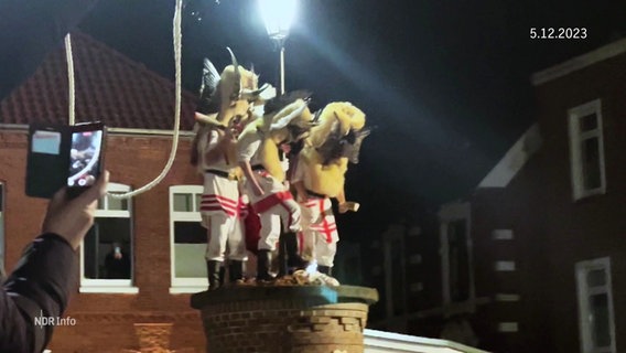 Als Klaasohm verkleidete Männer stehen auf einer Säule auf einem Borkumer Marktplatz bei Nacht. © Screenshot 