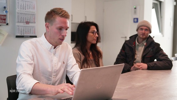 Ein Mann sitzt an einem Laptop, neben ihm sitzen zwei Personen. © Screenshot 