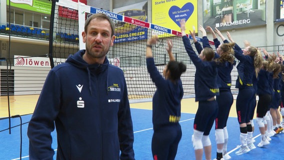 Felix Koslowski, Trainer vom SSC Palmberg Schwerin, blickt in die Kamera, während Volleyballspielerinnen am Netz das Blocken üben. © Screenshot 