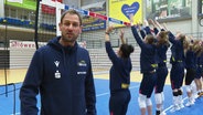 Felix Koslowski, Trainer vom SSC Palmberg Schwerin, blickt in die Kamera, während Volleyballspielerinnen am Netz das Blocken üben. © Screenshot 