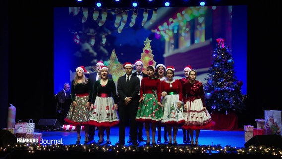 Auf einer weihnachtlich dekorierten Bühne stehen Männer und Frauen mit Weihnachtsmützen und singen. © Screenshot 