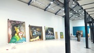 In einer Ausstellungsgallerie stehen größere, bunte Gemälde an einer Wand vor ihrer Montierung. © Screenshot 