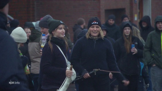 Zwei junge Frauen halten Kuhhörner in den Händen und nehmen freudig an einem Volksfest teil, im Hintergrund mehrere andere, junge Teilnehmende der Feierlichkeiten. © Screenshot 