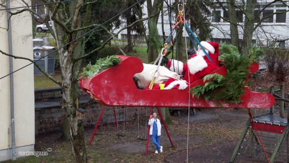 Ein Nikolaus in einem Holzschlitten, der durch Gurte gesichert mehrere Meter hoch in die Luft gezogen wird. © Screenshot 