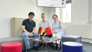 Zwei Männer und eine Frau sitzen hinter einer deutschen und chinesischen Flagge. © Screenshot 