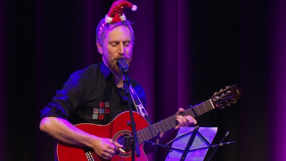 Ein Mann spielt auf einer Bühne Gitarre, er trägt eine Weihnachtsmann-Mütze. © Screenshot 