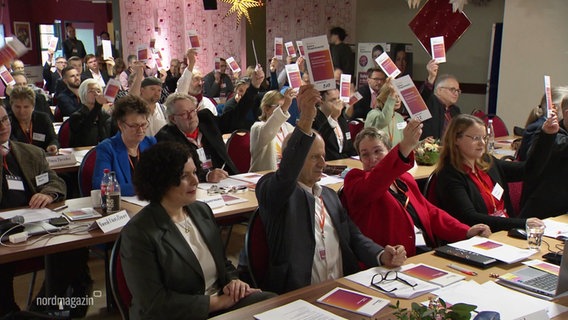 BSW Mitglieder sitzen in mehreren Stuhlreihen und heben ihren Stimmzettel. © Screenshot 