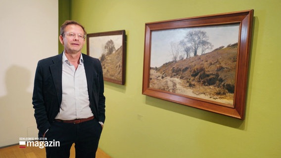 Florian Illies steht neben zwei Gemälden von Janus la Cour in der von ihm kuratierten Ausstellung auf Föhr. © Screenshot 