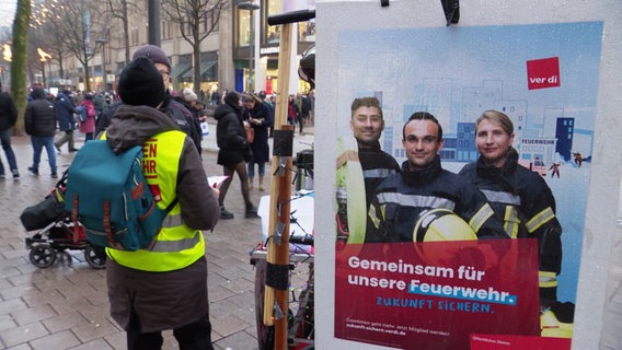 Ein Gewerkschaftsmitglied von ver.di steht in der Mönckebergstraße, die belebt ist mit Menschen, die dadurch schlendern. Daneben ein Plakat von ver.di, dass Solidarität mit der Hamburger Feuerwehr signalisiert. © Screenshot 