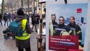 Ein Gewerkschaftsmitglied von ver.di steht in der Mönckebergstraße, die belebt ist mit Menschen, die dadurch schlendern. Daneben ein Plakat von ver.di, dass Solidarität mit der Hamburger Feuerwehr signalisiert. © Screenshot 