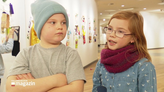 Zwei Kinder im Grundschulalter im Interview. Im Hintergrund sieht man einen Ausstellungsraum, in dem Bilder an den Wänden hängen. © Screenshot 