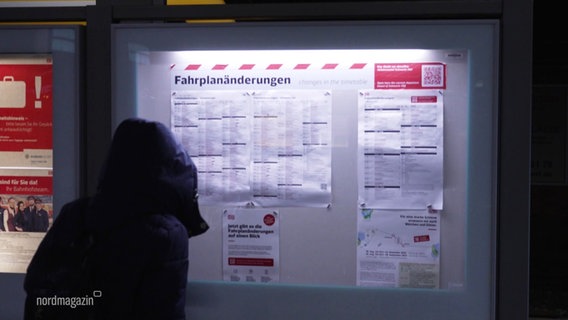 Eine Person mit dunkler Kapuzenjacke steht vor einer Vitrine mit ausgehängten Fahrplanänderungen an einem Bahnhof. © Screenshot 