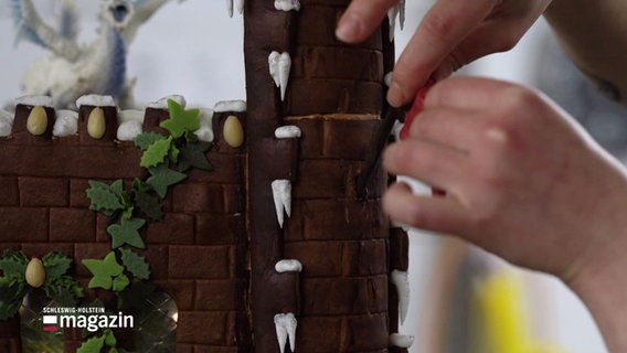 Jemand arbeitet an den letzten Details von einem Lebkuchenschloss. In den Teig sind Rillen, wie von Steinen, eingearbeitet, grünes Marzipan schlängelt sich als Efeu an dem Lebkuchengemäuer empor, Zuckerguss hängt als Eissazampfen herunter. © Screenshot 