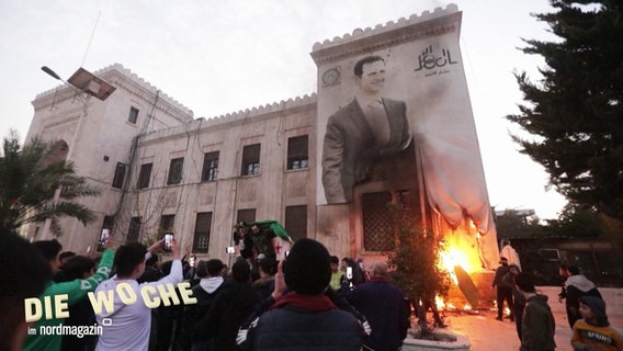 Menschen in Syrien haben sich vor einem Palast-ähnlichen Gebäude versammelt an dessen Fassade eine große Darstellung von Baschar al-Assad aufgemalt ist, vor dem Haus lodert ein Feuer. © Screenshot 