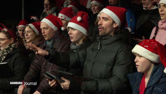 Eine Gruppe von Menschen mit Weihnachtsmützen singt auf einer Bühne. © Screenshot 