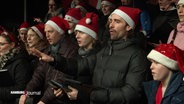 Eine Gruppe von Menschen mit Weihnachtsmützen singt auf einer Bühne. © Screenshot 