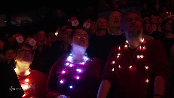 Menschen mit Lichterketten und Weihnachtsmann-Motiven als Kopfschmuck. © Screenshot 