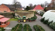 Auf einem Hof stehen Weihnachtsbäume zum Verkauf, ein Traktor zieht einen Anhänger mit Tannenbäumen. © Screenshot 