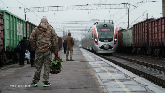 Ein Soldat in Uniform steht mit einem Strauß roter Rosen auf einem Bahnsteigu nd wartet auf einen einfahrenden Zug. © Screenshot 