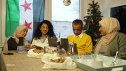 Eine syrische Familie sitzt am Tisch, im Hintergrund ein Weihnachtsbaum, am Fenster hängt die syrische Flagge. © Screenshot 