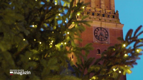 Ein Ausschnitt eines Tannenbaums mit Lichtern, im Hintergrund der Turm des historischen Rathauses von Kiel. © Screenshot 