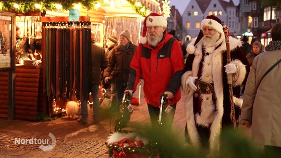 Zwei Weihnachtsmänner, einer am Rollator, laufen über einen Weihnachtsmarkt. © Screenshot 