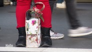 Am Bahnsteig: Zwischen den Füßen einer Person in roter Hose steht eine Tüte mit Geschenken. © Screenshot 