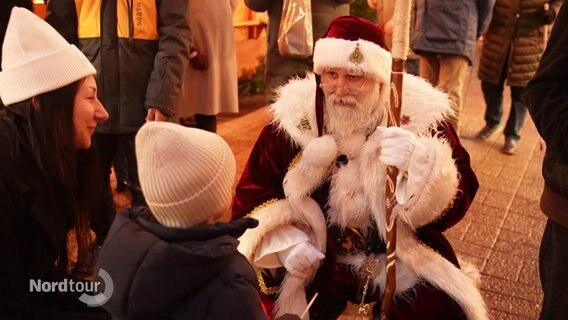 Ein Weihnachtsmann kniet auf einem Weihnachtsmann vor einem Jungen um mit ihm auf Augenhöhe zu sprechen. © Screenshot 