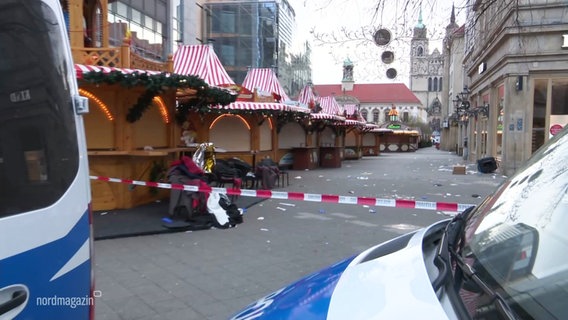 Auf einem Weihnachtsmarkt sind tagsüber alle Buden geschlossen, Absperrband hängt zwischen zwei Einsatzwagen der Polizei vor dem Eingang zum Weihnachtsmarkt. © Screenshot 