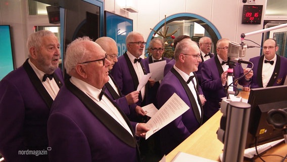 Ein älterer Männerchor, tgekleidet in schicken, lila Anzügen, singt in einem Radiostudio. © Screenshot 