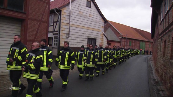 Viele Einsatzkräfte der Feuerwehr gehen in einem Trauerzug durch eine Dorfstraße. © Screenshot 