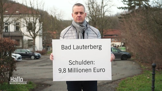 Der Bürgermeister von Bad Lauterberg hälz ein Schild hoch auf dem die Schulden der gemeinde zusehen sind. © Screenshot 