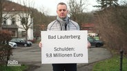 Der Bürgermeister von Bad Lauterberg hälz ein Schild hoch auf dem die Schulden der gemeinde zusehen sind. © Screenshot 