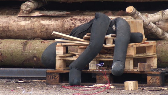 Eine gepolsterte Puppen-Attrappe liegt nach hinten umgekippt auf einem Stapel Paletten nach einem Feuerwerkskörper-Test. © Screenshot 