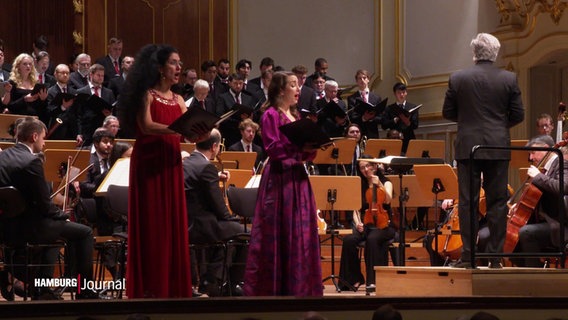 Zwei Personen in Kleidern singen vor einem Orchester in einem Konzertsaal. © Screenshot 