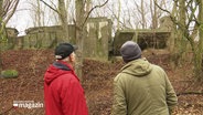 Zwei Männer schauen auf eine Ruine im Wald. © Screenshot 