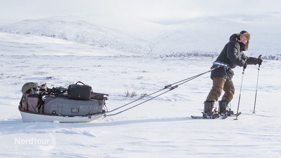 Naturfotograf Mario Müller stapft mit Schneeschuhen durch eine Winterlandschaft und zieht einen Schlitten mit Ausrüstung hinter sich her. © Screenshot 