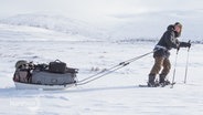 Naturfotograf Mario Müller stapft mit Schneeschuhen durch eine Winterlandschaft und zieht einen Schlitten mit Ausrüstung hinter sich her. © Screenshot 