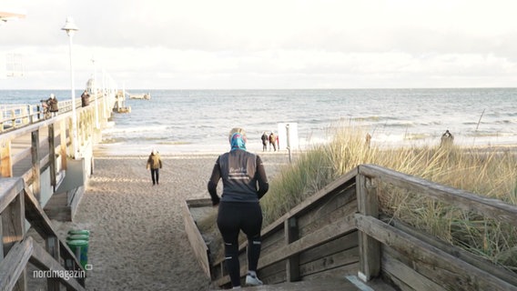 Meschen gehen am Meer spazieren, eine Joggerin läuft eine Treppe zum Strand hinunter. © Screenshot 