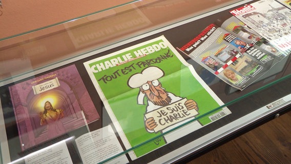 Eine Ausgabe des Satiremagazins "Charlie Hebdo" liegt in einer Vitrine. © Screenshot 