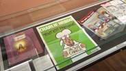 Eine Ausgabe des Satiremagazins "Charlie Hebdo" liegt in einer Vitrine. © Screenshot 