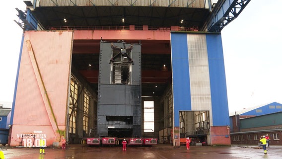 Ein riesiges Schleusentor wird aus der Montagehalle transportiert - die Arbeiter im Vordergrund wirken dagegen winzig klein. © Screenshot 