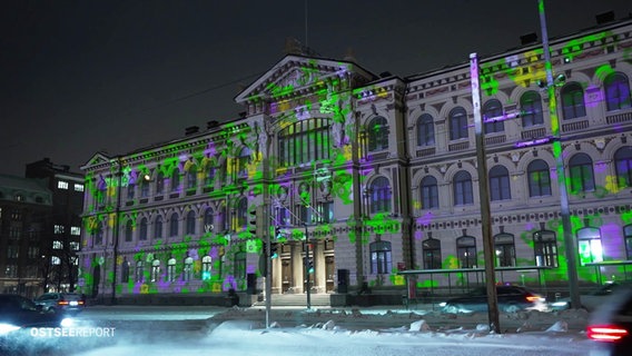 Ein größeres, palastähnliches Staatsgebäude ist bei Nacht von bunten Lichtern angestrahlt. © Screenshot 